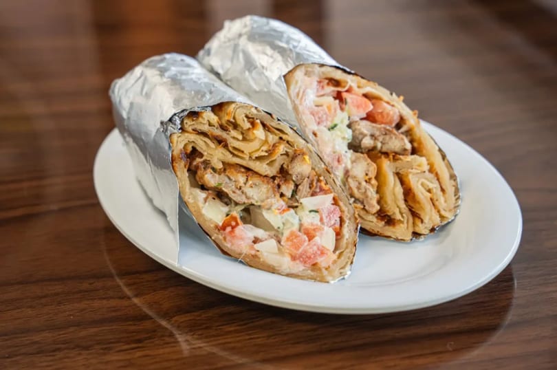 Two foil-wrapped shawarma wraps, cut in half, revealing chicken, tomatoes, and sauce on a white plate.