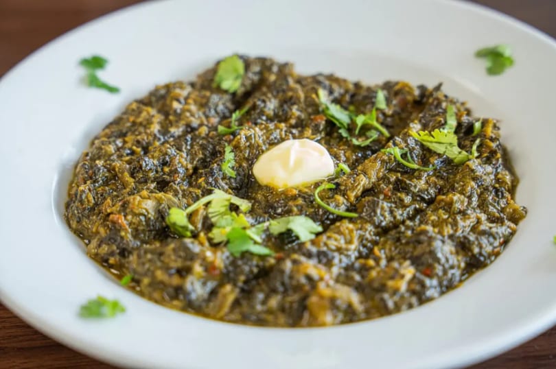 A white bowl filled with green saag, garnished with cilantro and a dollop of butter.