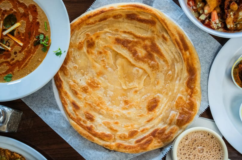 Golden, flaky paratha bread on a white plate, surrounded by various Indian dishes and a cup of chai.