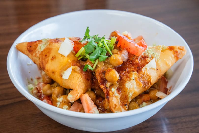 Samosa chaat with chickpeas, tomatoes, and cilantro in a white bowl on a wooden table.