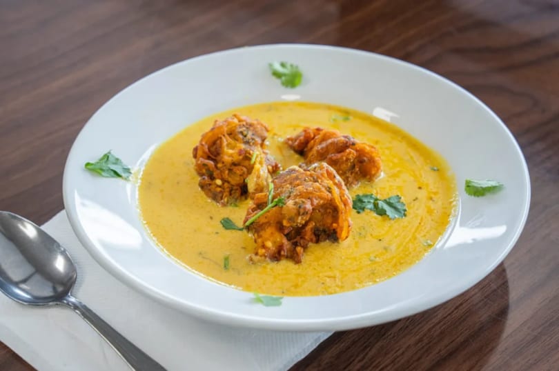 Three pakoras in a yellow curry sauce, garnished with cilantro, on a white plate with a spoon.